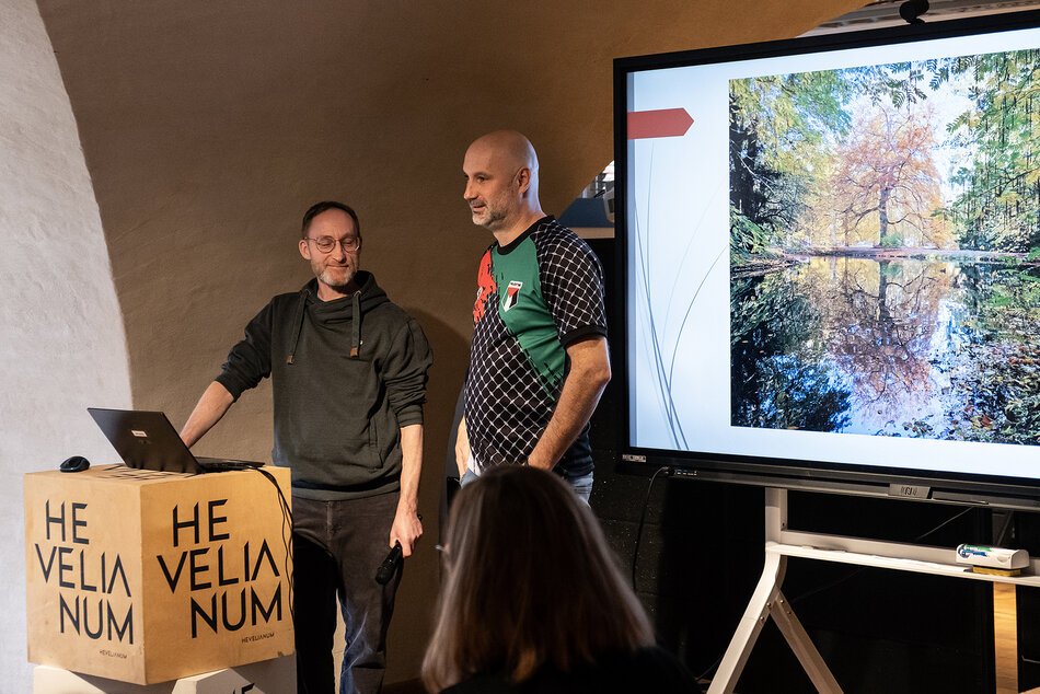 Dwóch mężczyzn stoi na mównicy w sali konferencyjnej, obok stoi ekran.