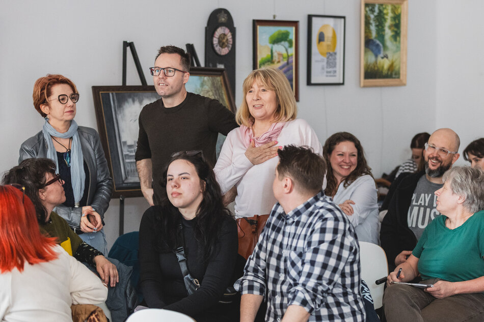 na zdjęciu kilka osób część siedzi na krzesłach, trzy osoby stoją patrzą przed siebie jakby z kimś rozmawiały przed nimi