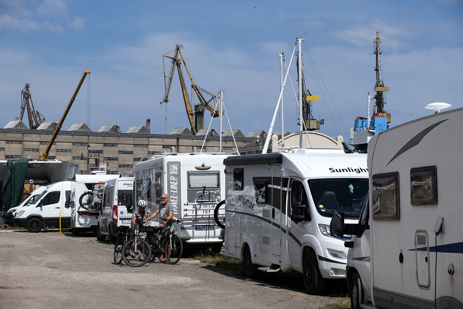 Rząd kamperów zaparkowanych na terenie stoczni, w tle hala stoczniowa i dźwigi. Przy kamperze dwie osoby z rowerami.