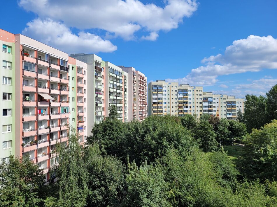 Zdjęcie przedstawia osiedle wielopiętrowych bloków mieszkalnych o kolorowych elewacjach, otoczonych gęstą, zieloną roślinnością. Budynki charakteryzują się licznymi balkonami i są ustawione w rzędach pod pogodnym, błękitnym niebem z białymi chmurami.