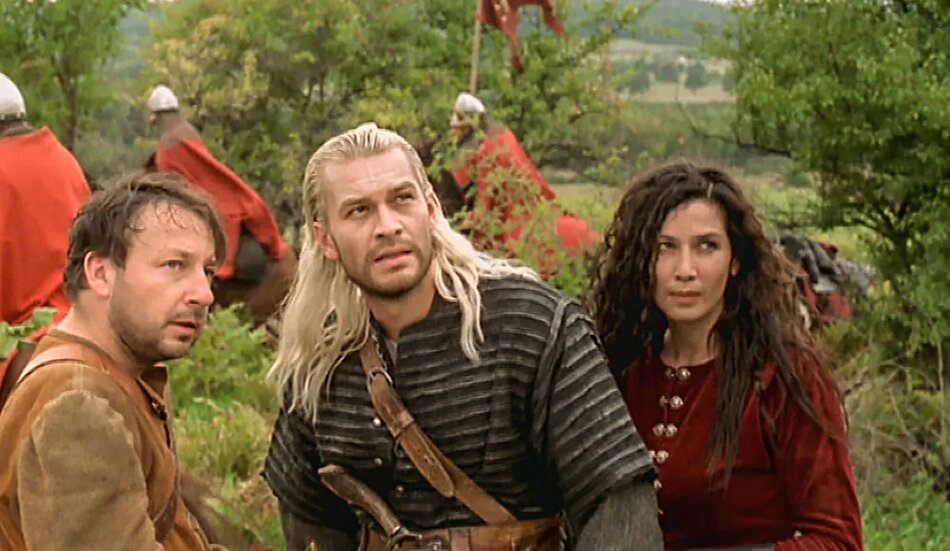 Kadr z filmu Wiedźmin. Kobieta i dwóch mężczyzn na pierwszym planie patrzą w jednym kierunku, ubrani są w stroje ze średniowiecza, jeden ma długie blond włosy i kolczugę. W tle rycerze w czerwonych płaszczach i hełmach na głowach. Za nimi drzewa i krzaki.