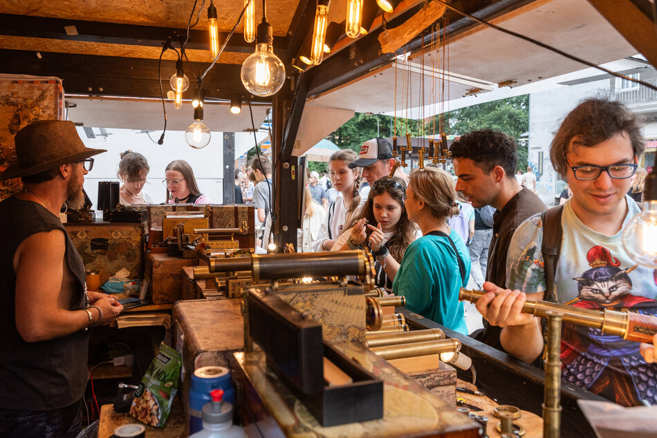 Zdjęcie przedstawia tętniące życiem stoisko rzemieślnicze w stylu steampunk, wypełnione mosiężnymi lunetami i dekoracyjnymi mechanizmami. Wokół lady gromadzą się ludzie, którzy z zaciekawieniem oglądają wystawione przedmioty pod ciepłym światłem dekoracyjnych żarówek.