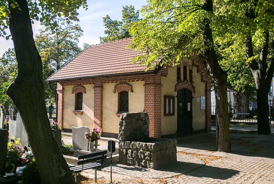 na zdjeciu kamienna studnia, w tle niewysoki budynek o jasnej elewacji