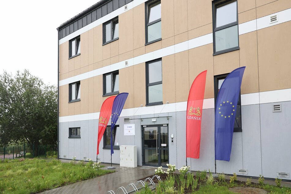 na zdjęciu budynek nowoczesny o pomarańczowej elewacji, przed drzwiami wejściowymi flagi czerwone Gdańska i niebieskie unii europejskiej