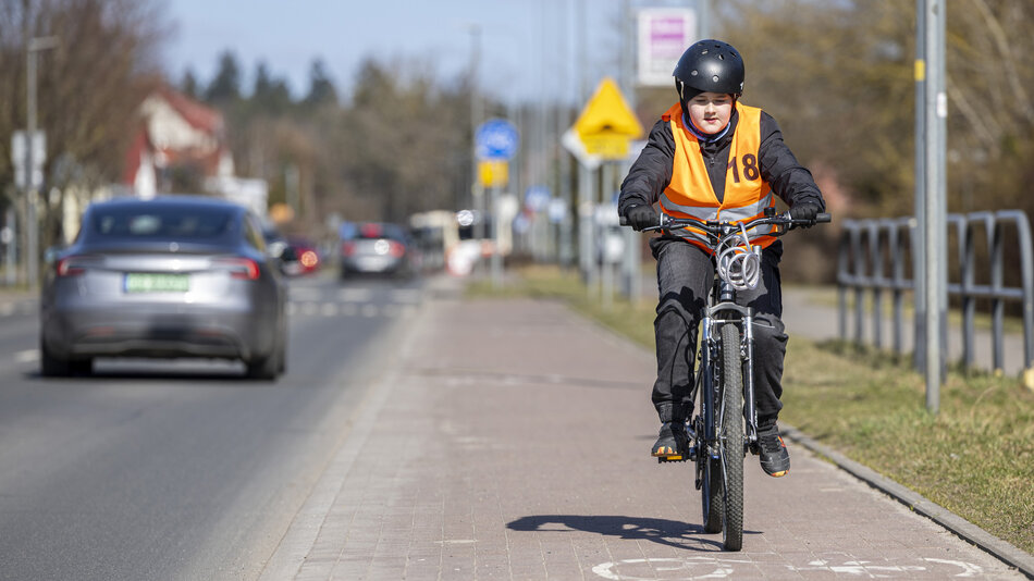 Zdjęcie przedstawia młodego rowerzystę w kasku i odblaskowej kamizelce, który porusza się wyznaczoną ścieżką rowerową wzdłuż jezdni. W tle widać jadące samochody oraz infrastrukturę miejską, co podkreśla znaczenie bezpieczeństwa w ruchu drogowym.