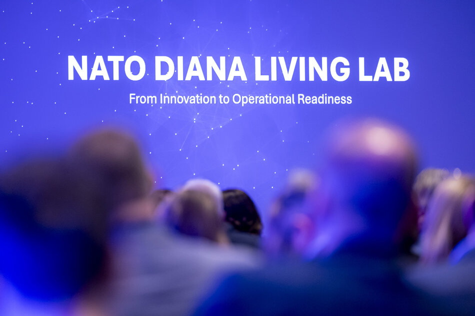 Na grafice widoczny jest duży napis „NATO DIANA LIVING LAB” wraz z hasłem o przechodzeniu od innowacji do gotowości operacyjnej. Napis znajduje się na niebieskim tle z motywem siatki połączeń, a na pierwszym planie widać rozmyte sylwetki osób uczestniczących w wydarzeniu.