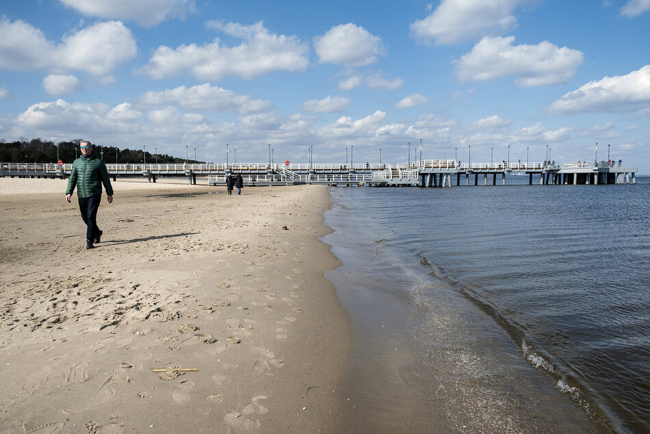 Styk morza i plaży, po której idzie mężczyzna. W tle widać molo.