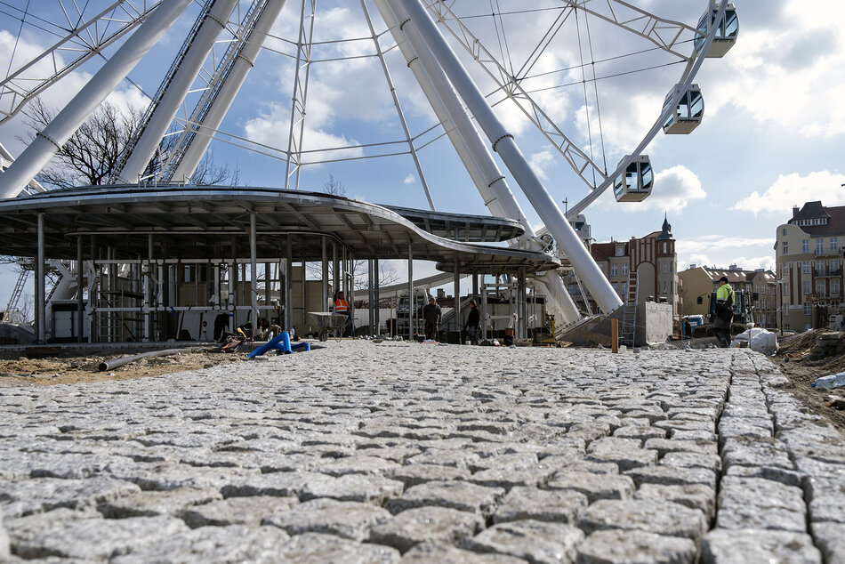 Prace budowlane przy kole widokowym, na pierwszym planie układana kostka brukowa, w tle stalowe konstrukcje z zadaszeniami i same koło widziane do jednej trzeciej konstrukcji.
