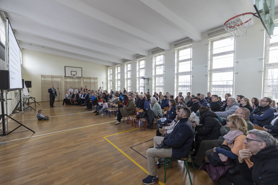 szkolna sala gimnastyczna wypełniona ludźmi, ponad sto osób siedzi na krzesłach, patrzy przed siebie