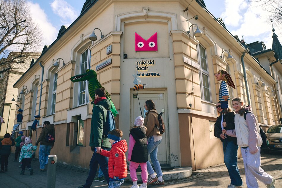Zdjęcie przedstawia wejście do Miejskiego Teatru Miniatura, przed którym spacerują dorośli i dzieci w słoneczny dzień. Na elewacji budynku widoczne są charakterystyczne dekoracje, w tym duża figura zielonego krokodyla oraz logo teatru z sową. 