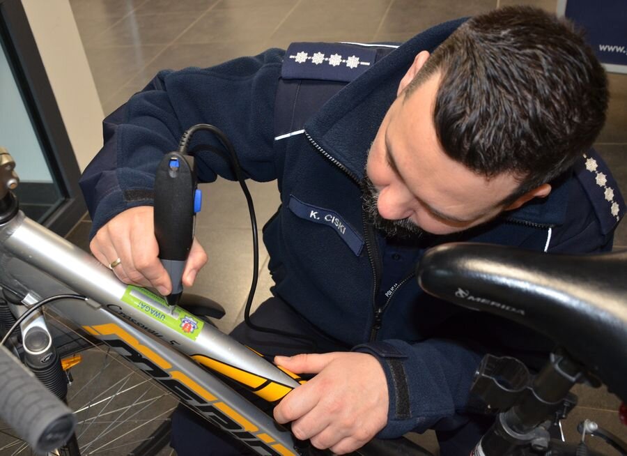 Zdjęcie przedstawia funkcjonariusza policji, który za pomocą grawera nanosi numer identyfikacyjny na ramę roweru. Akcja ta ma na celu zabezpieczenie mienia przed kradzieżą i ułatwienie jego późniejszej identyfikacji przez służby.