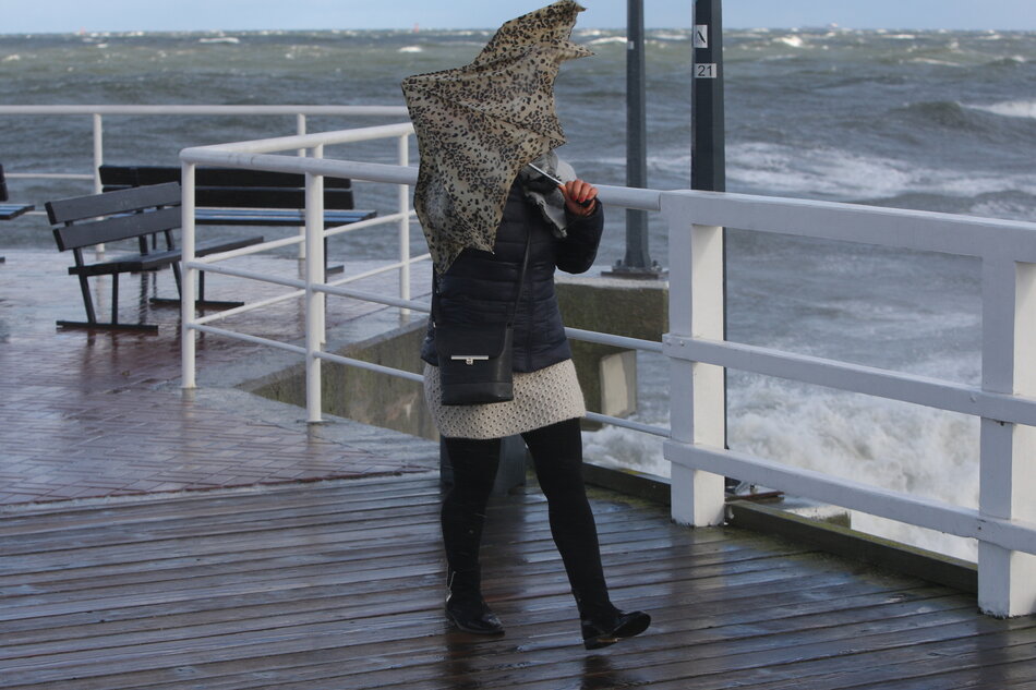 Kobieta podczas wichury na molo w Brzeźnie. Widać wzburzone morze, kobieta trzyma nad głową parasolkę wyginaną przez wiatr