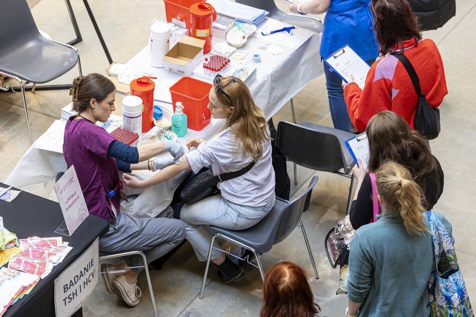 Grupa ludzi zebranych wokół stołu w pokoju. Na zdjęciu cztery osoby, trzy kobiety i jeden mężczyzna. Stół jest pokryty białym obrusem i znajdują się na nim różne przedmioty, w tym butelki, pudełka i inne artykuły medyczne. Kobieta po lewej stronie nosi purpurową koszulę i wydaje się być pielęgniarką lub lekarzem. Siedzi na krześle i ma w ręku schowek i długopis. Obok niej jest inna kobieta w pomarańczowej kurtce i noszący stetoskop wokół szyi. Po prawej stronie również trzyma schowek. Przed nimi można zobaczyć kilka innych osób siedzących na ławkach i przyglądających się scenie.