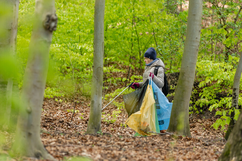 Człowiek w kurtce i czapce idzie przez las i niesie worki na śmieci. W ręku trzyma szczypce do podnoszenia z ziemi śmieci.