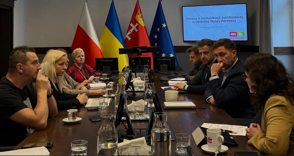 zdjęcie ze spotkania, po obu stronach dużego ciemnego stołu siedzą dorosłe osoby, rozmawiają ze sobą