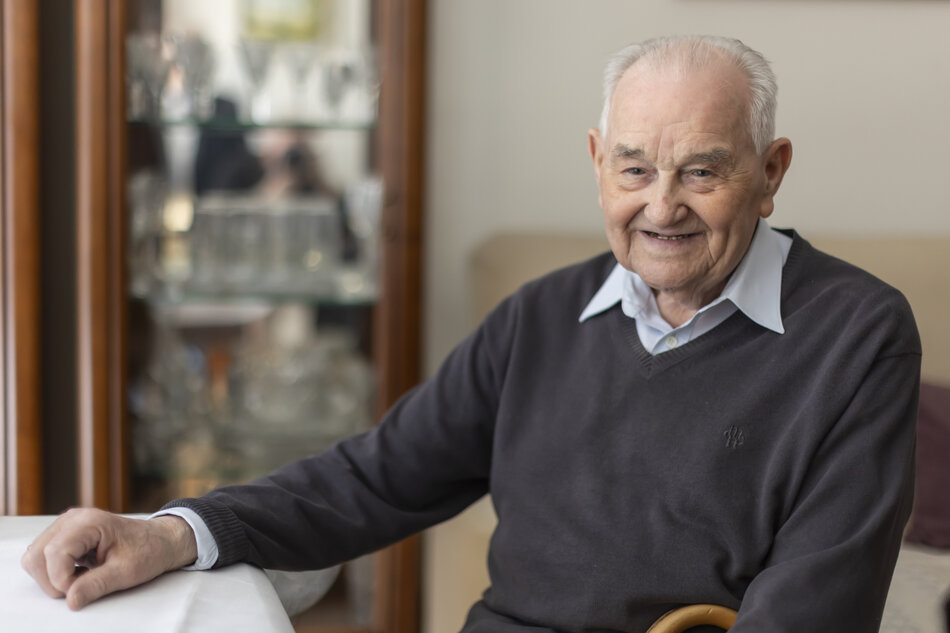 Starszy mężczyzna pozuje do zdjęcia w mieszkaniu siedząc za stołem, uśmiecha się.