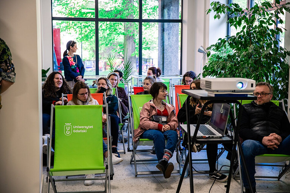 W sali siedzą ludzie na leżakach, przed nimi stoi projektor na stoliku.