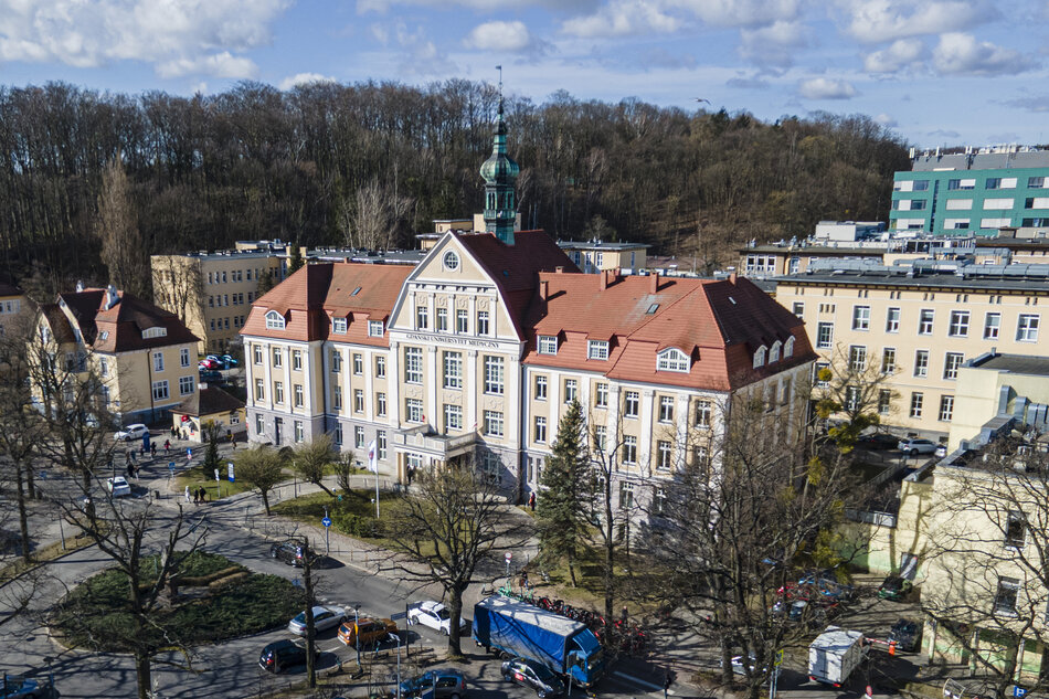 Zdjęcie przedstawia zabytkowy gmach główny Gdańskiego Uniwersytetu Medycznego z charakterystyczną wieżyczką i czerwonym dachem. Budynek otoczony jest nowoczesną infrastrukturą szpitalną oraz drzewami, a w tle widoczne jest zalesione wzgórze.