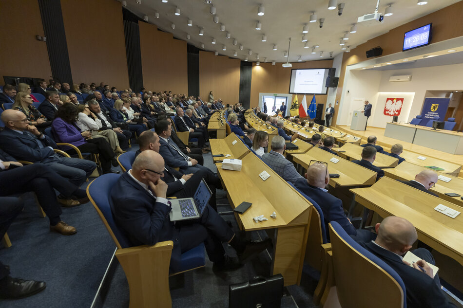 W sali obrad siedzi kilkadziesiąt osób. Na mównicy przemawia mężczyzna.