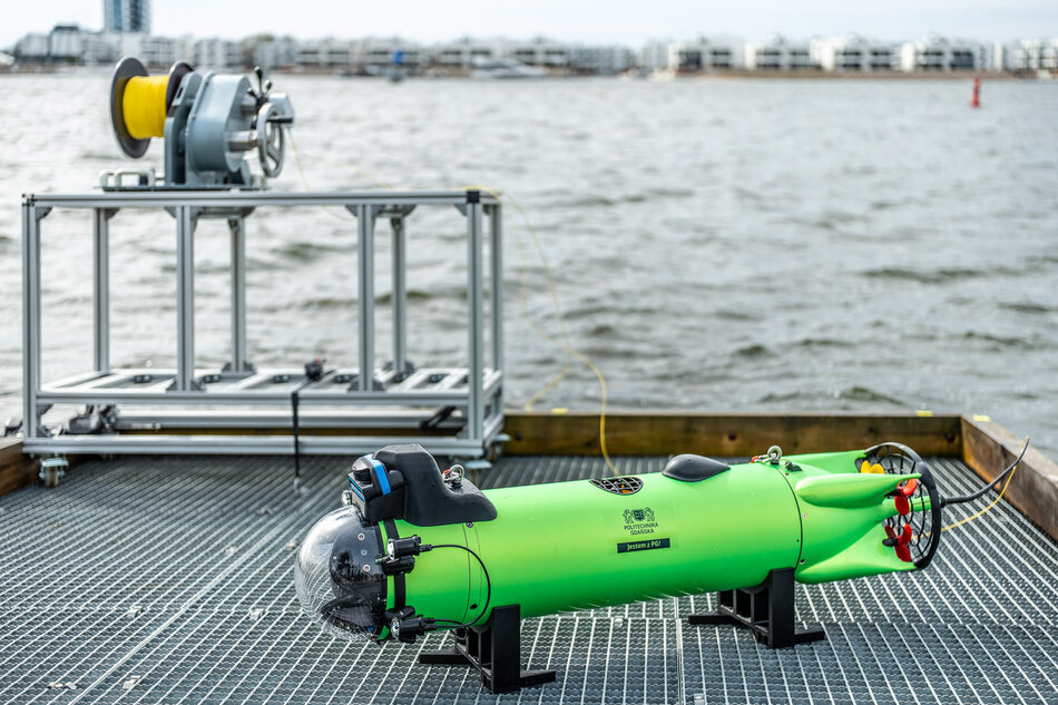 Na kratowanym pomoście spoczywa jaskrawozielony, podłużny dron podwodny wyposażony w przezroczystą kopułę kamery i śruby napędowe. W tle, obok metalowego stelaża z wyciągarką, rozciąga się widok na lekko wzburzoną wodę oraz zabudowania na drugim brzegu.