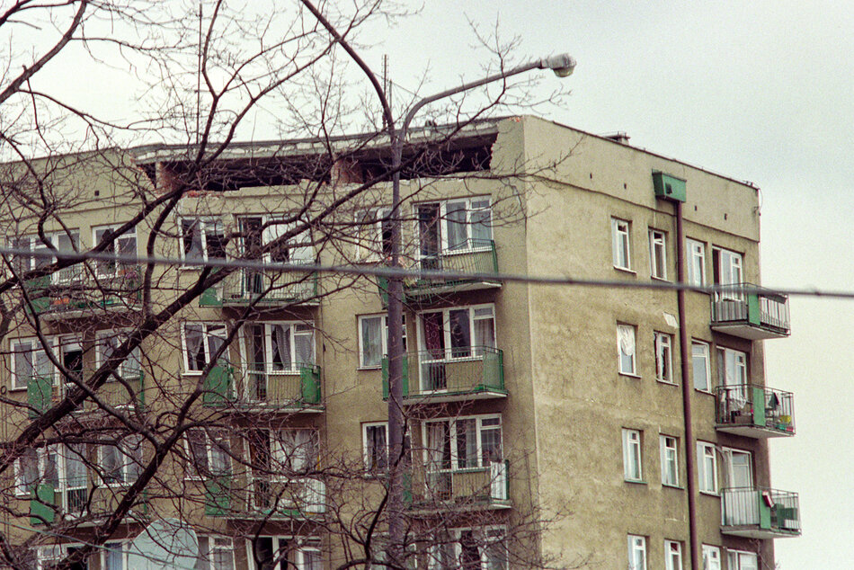 Zdjęcie przedstawia zniszczony blok mieszkalny z wyraźną wyrwą w górnej części elewacji oraz powybijanymi szybami w oknach. Widoczne w pobliżu bezlistne gałęzie drzew i ponura kolorystyka nadają scenie surowy, dramatyczny charakter. 
