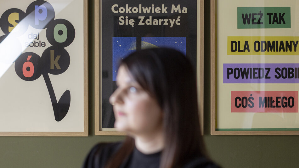 Na pierwszym planie widoczna jest nieostra sylwetka kobiety, która stoi na tle ściany z trzema oprawionymi plakatami z polskimi napisami. Grafiki zawierają motywacyjne hasła, takie jak „daj sobie spokój” czy „powiedz sobie coś miłego”, przedstawione w nowoczesnej i kolorowej formie. 