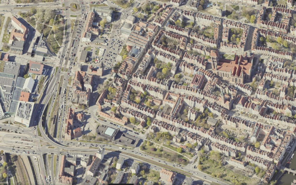 Zdjęcie przedstawia widok z lotu ptaka na gęsto zabudowane centrum miasta z charakterystycznymi kamienicami o czerwonych dachach oraz dużym, ceglanym kościołem. W lewej części kadru widoczna jest rozbudowana infrastruktura drogowa z wielopasmowymi jezdniami, torowiskami i nowoczesnymi budynkami użytkowymi.