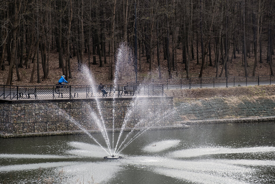 Fontanna na zbiorniku retencyjnym. Na jego brzegu siedzi osoba na ławce, obok jedzie osoba na rowerze.