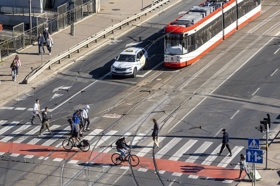 Zdjęcie przedstawia ruchliwe skrzyżowanie w mieście, na którym widać biało-czerwony tramwaj, jasny samochód osobowy oraz licznych pieszych i rowerzystów korzystających z pasów. Całość ujęta jest z perspektywy lotu ptaka, co pozwala dostrzec skomplikowaną sieć trakcyjną oraz wyraźne, czerwone oznaczenia drogi rowerowej na asfalcie.
