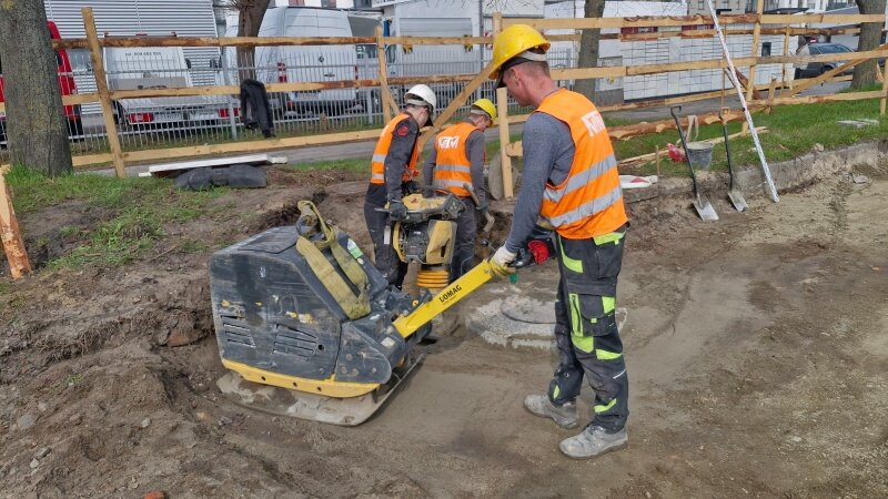 na zdjęciu dwóch robotników w roboczych pomarańczowych kamizelkach i kaskach, jeden trzyma maszynę do utwardzania gleby