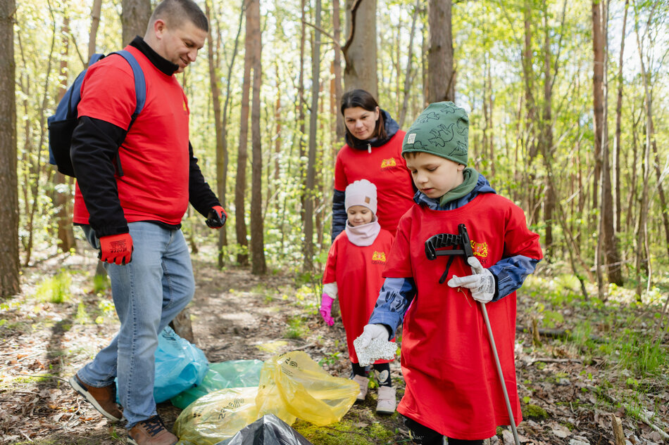 Zdjęcie przedstawia czteroosobową rodzinę, która wspólnie sprząta las, zbierając śmieci do kolorowych worków. Wszyscy są ubrani w czerwone koszulki i ochronne rękawiczki, a najmłodszy chłopiec z zaangażowaniem pomaga dorosłym, używając specjalnego chwytaka.