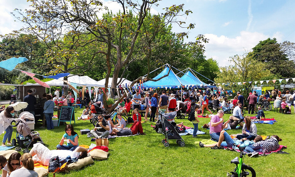 Ludzie na dworze. Dookoła zielono. Widać kolorowe stoiska.