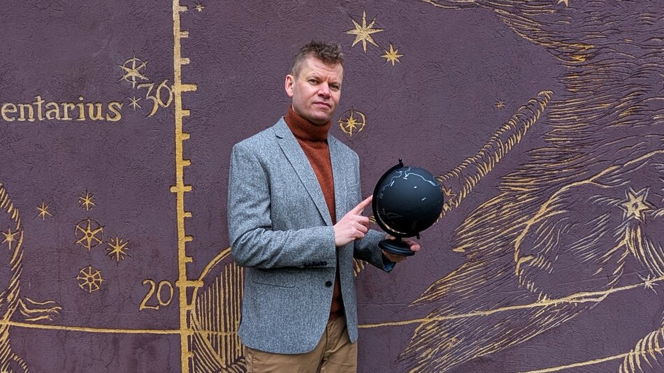 Osoba stoi na tle ściany ozdobionej ilustracją z motywami astronomicznymi, takimi jak gwiazdy, linie i oznaczenia przypominające mapę nieba. W dłoniach trzyma czarny globus, wskazując na niego palcem. Osoba ubrana jest w marynarkę i golf, a cała scena znajduje się na zewnątrz, przy dekoracyjnej elewacji budynku.