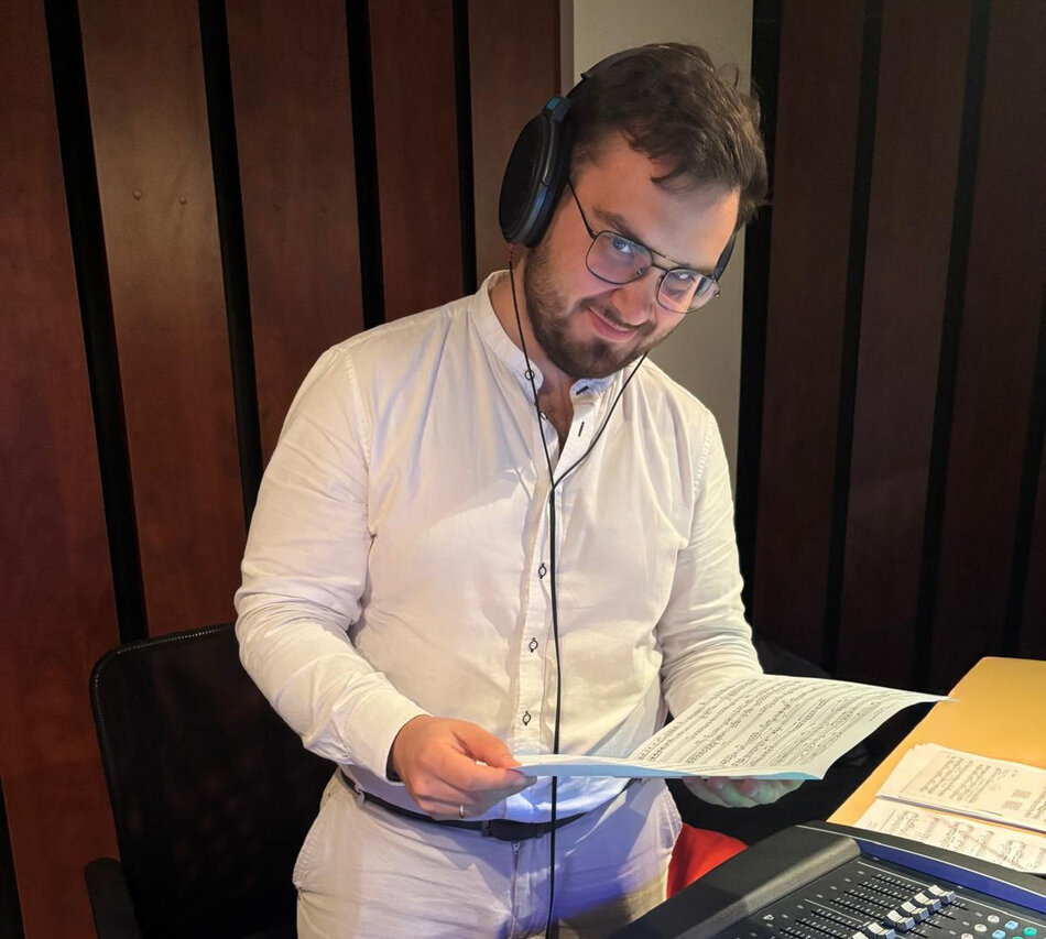 Osoba stoi przy pulpicie z urządzeniami audio i trzyma w dłoniach kartki z nutami lub tekstem. Na uszach ma słuchawki połączone przewodem, a na blacie widoczne są mikser dźwięku oraz dokumenty. Osoba ubrana jest w jasną koszulę. W tle znajduje się ściana z pionowymi panelami akustycznymi, sugerująca przestrzeń studia nagraniowego lub reżyserskiego.