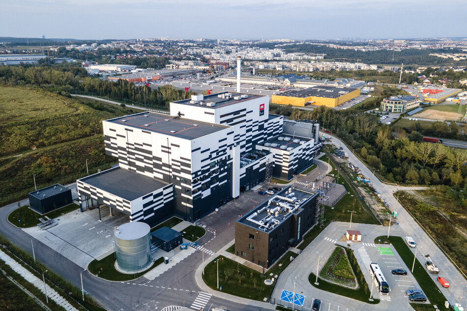 Zdjęcie przedstawia nowoczesny, wielkogabarytowy budynek przemysłowy o biało-czarnej elewacji, który jest siedzibą Portu Czystej Energii w Gdańsku. Obiekt otoczony jest infrastrukturą drogową i parkingami, a w oddali rozciąga się panorama miasta z licznymi zabudowaniami i terenami zielonymi.