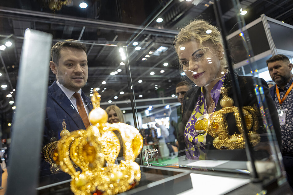 Na zdjęciu widać bogato zdobioną, złotą koronę z elementami bursztynu, która jest eksponowana w szklanej gablocie podczas targów bursztynu Amberif