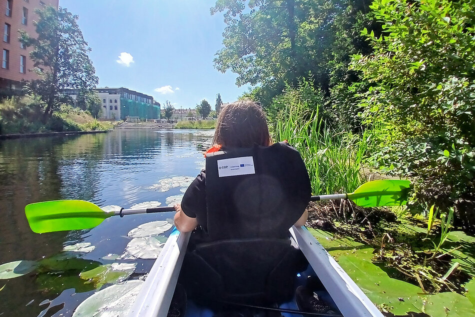 Zdjęcie zrobione z kajaku. Na pierwszym planie plecy osoby z wiosłami siedzącej w kajaku na rzece w mieście. Wokół drzewa i budynki.