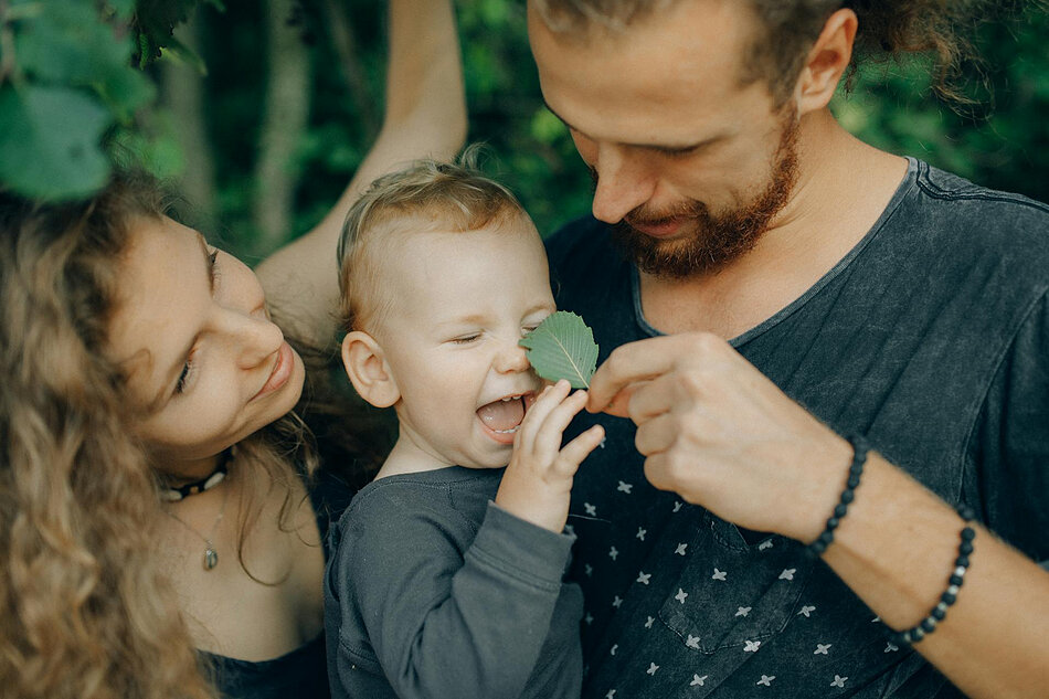 Młodzi kobieta i mężczyzna z dzieckiem pośród zieleni. Mężczyzna dotyka nosa dziecka liściem, te się śmieje.