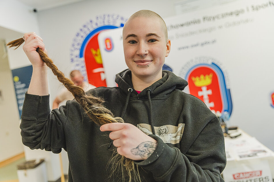 Łysa dziewczyna trzyma ścięty warkocz włosów i uśmiecha się do kamery