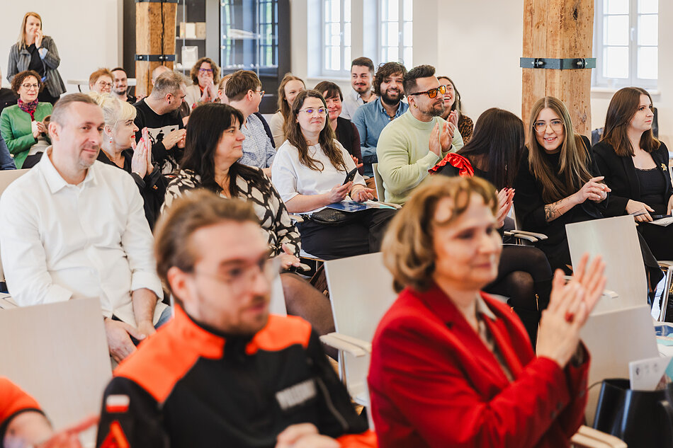 Na zdjęciu widać grupę uśmiechniętych osób siedzących w rzędach i oklaskujących kogoś znajdującego się poza kadrem. Uczestnicy są ubrani w różnorodne stroje, od codziennych ubrań po charakterystyczny czarno-pomarańczowy mundur, co wskazuje na oficjalny charakter tego wydarzenia. 