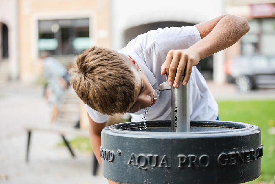 Zdjęcie przedstawia chłopca pijącego wodę bezpośrednio z nowoczesnego poidełka miejskiego z napisem „tap water”. Urządzenie z hasłem „AQUA PRO GENERATION” jest przykładem ogólnodostępnego punktu z darmową wodą pitną w przestrzeni publicznej. 