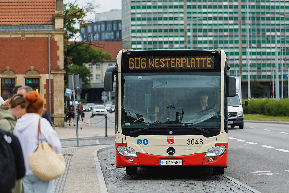 Zdjęcie przedstawia miejski autobus marki Mercedes-Benz o numerze 3048, który obsługuje linię 606 kierującą się w stronę Westerplatte. Pojazd w gdańskich barwach stoi na przystanku, a w tle widać miejską zabudowę oraz pasażerów oczekujących na wejście do środka.