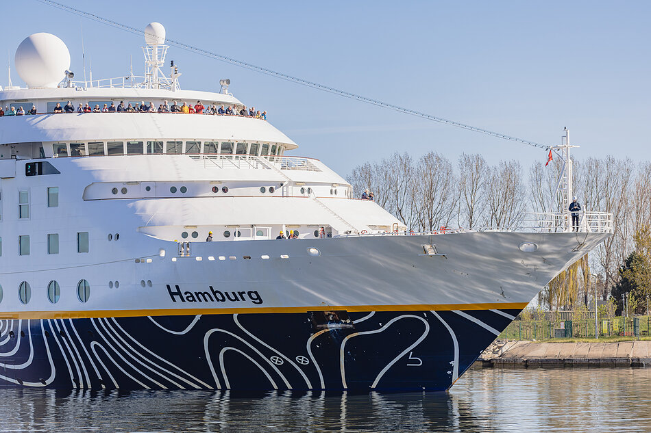 Zdjęcie przedstawia duży statek wycieczkowy o nazwie „Hamburg”, płynący po spokojnej wodzie na tle bezchmurnego nieba. Jego biały kadłub zdobią charakterystyczne ciemnoniebieskie wzory, a na górnych pokładach można dostrzec licznych pasażerów obserwujących okolicę.