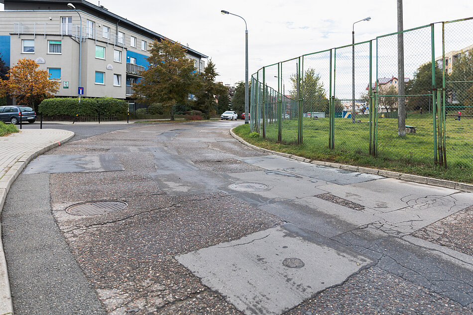 zdjęcie nawierzchni ulicy asfaltowej, która jest zniszczona, w tle blok mieszkaniowy