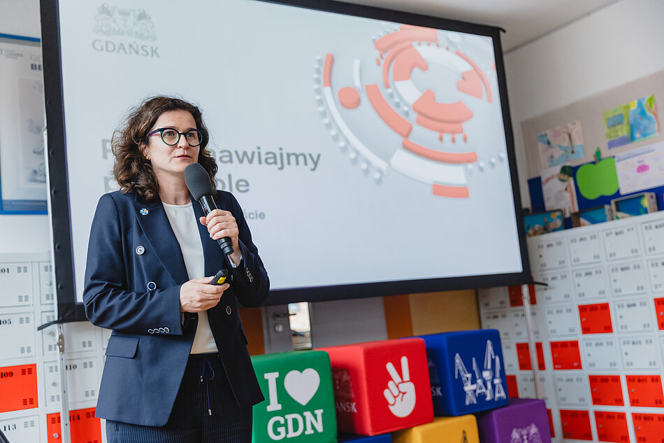 na zdjęciu prezydent gdańska w granatowej garsonce, mówi do mikrofonu, za nią wyświetla się kolorowy baner