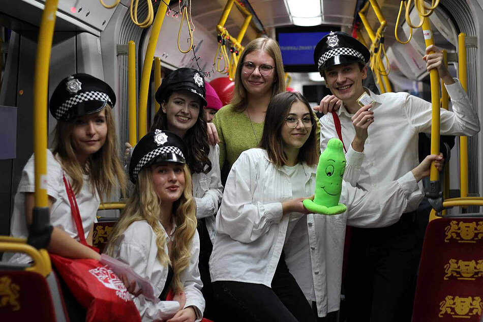 Grupa młodych ludzi w czapkach policyjnych pozuje do zdjęcia w tramwaju. Jedna z nich trzyma w rękach maskotkę prezerwatywy. Wszyscy się śmieją.