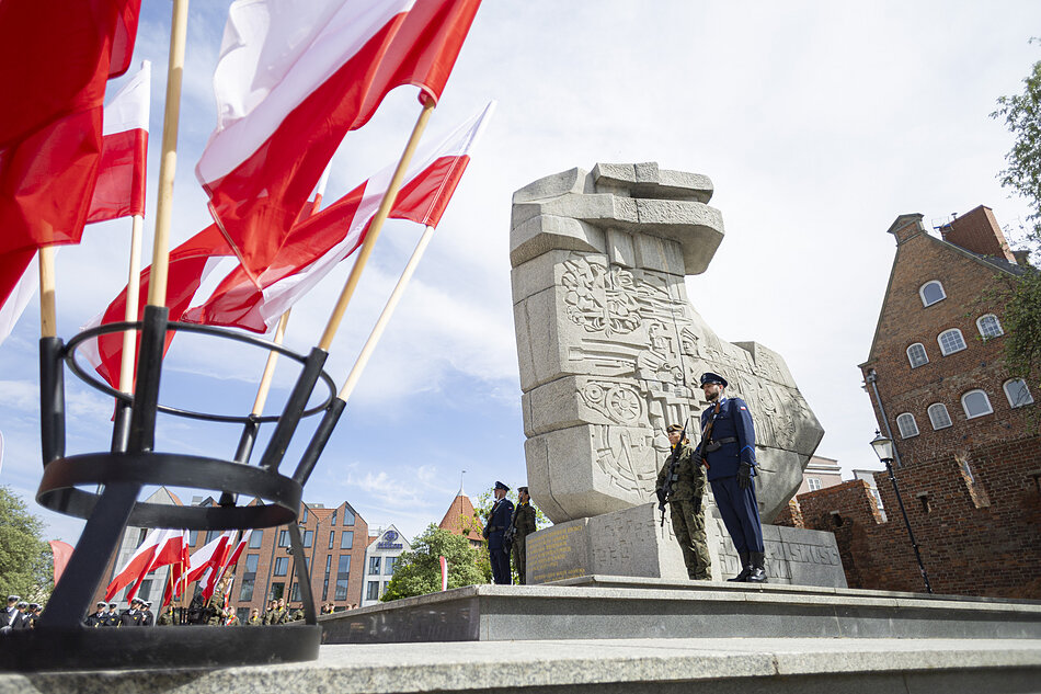 na zdjęciu w tle jasny wysoki pomnik przy którym stoi żołnierz, po lewej widać kilka stojących biało czerwonych flag