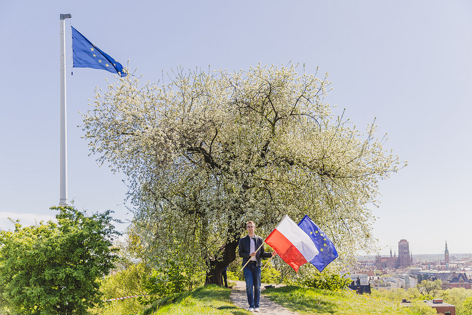 Flaga Europy powiewa na wysokim maszcie na górze nad starym miastem. Obok masztu kwitnące drzewko owocowe, pod którym idzie młody człowiek z flagami Polski i Europy.
