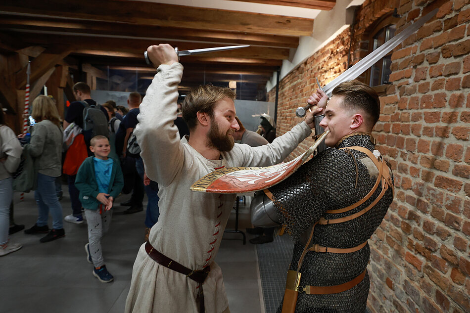 Zdjęcie przedstawia dwóch mężczyzn biorących udział w pozorowanej walce średniowiecznej, z których jeden ma na sobie kolczugę, a drugi białą tunikę. W tle widać grupę zwiedzających, co sugeruje, że pokaz odbywa się w muzeum lub podczas historycznego festynu.