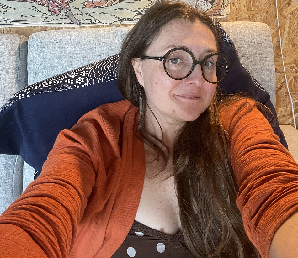 Kolorowe zdjęcie typu selfie przedstawiające osobę siedzącą na kanapie w pomieszczeniu. Osoba znajduje się blisko aparatu i patrzy w obiektyw, ma na sobie duże, okrągłe okulary oraz długie kolczyki. Ubrana jest w pomarańczowy sweter, a spod niego widoczna jest część innego elementu garderoby w kropki. W tle znajduje się jasna tapicerka kanapy, ciemnoniebieska poduszka z jasnym wzorem oraz ściana z materiału o charakterystycznej fakturze, na której zawieszony jest fragment ilustracji lub obrazu.
